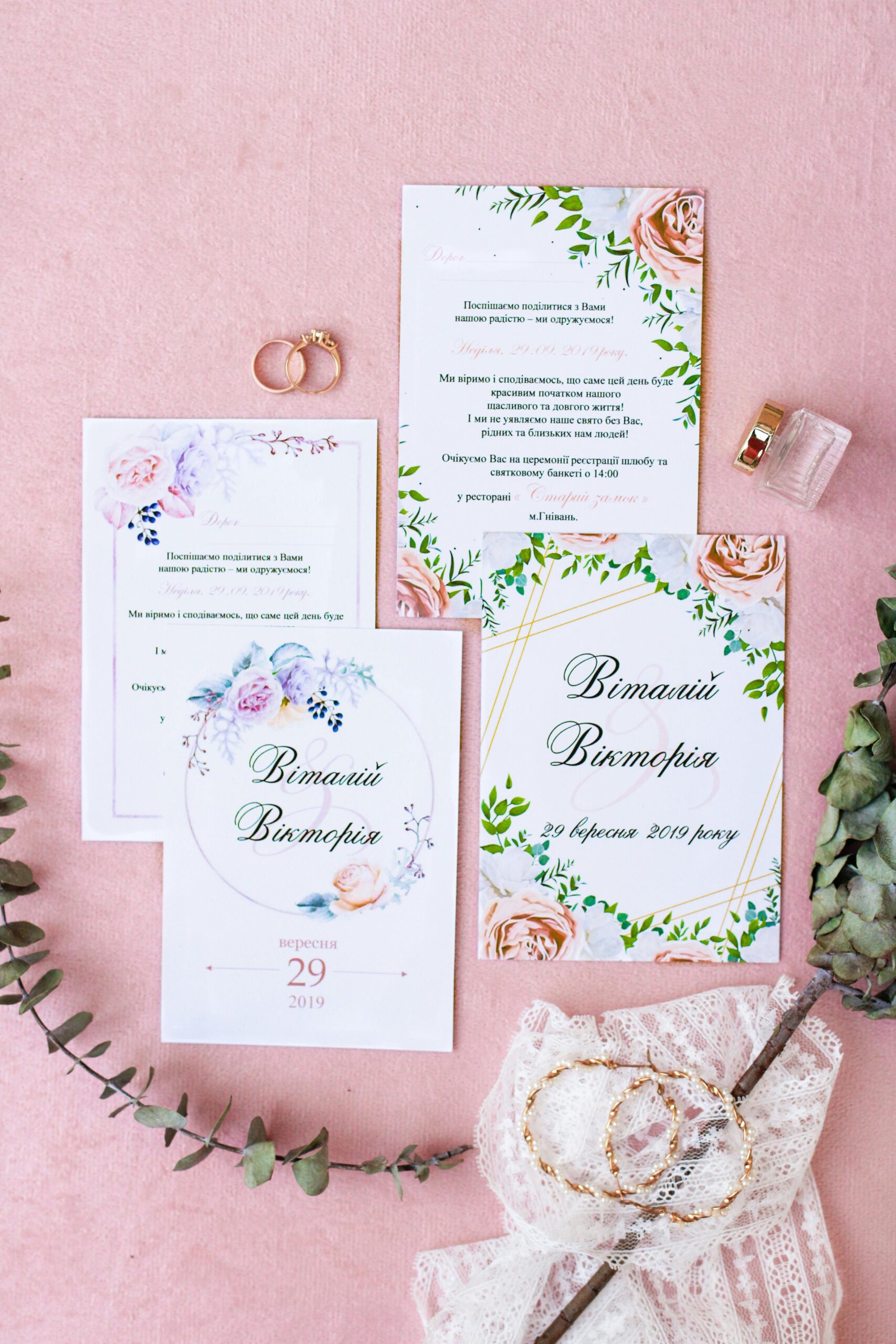 Chic floral wedding invitations flat lay with lace, rings, and perfume on a pink background.